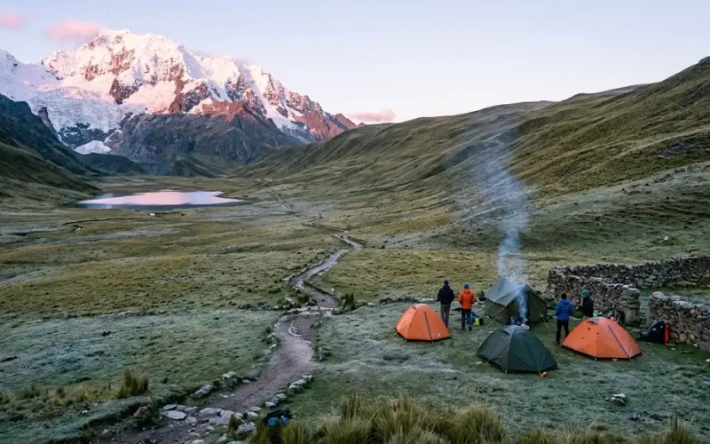 Campamento de tiendas junto a un valle andino con un nevado al fondo y un sendero que lleva a otras rutas de trekking en Perú